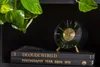 A black alarm clock with gold accents sits on top of two black books on a shelf, with a green plant in the upper left corner. Anteprima dell’immagine prodotto