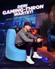A man is sitting on a light blue beanbag chair in a gaming room, holding a game controller. The room has a city skyline view through a window and gaming equipment. Produktbild-Vorschau