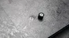 A close-up view of a gray textured surface with two small metallic and black components, possibly part of a table mechanism. Produktbild-Vorschau