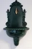 A dark green, ornate wall-mounted fountain with a brass faucet is displayed against a white brick wall. Produktbild-Vorschau