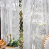 A close-up view of white lace curtains with a floral embroidery pattern, revealing a glimpse of trees and foliage through a wavy opening in the center. A table setting with bread and candles is visible in the foreground. Náhľad obrázku produktu