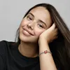 A young woman with freckles smiles as she rests her head on her hand, showing off a bracelet with a red heart charm. Produktbild-Vorschau
