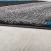 A close-up view of the edge of a modern rug with geometric patterns in turquoise, gray, and black, resting on a light-colored wooden floor. Anteprima dell’immagine prodotto