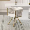 A beige upholstered chair with gold legs sits on a patterned rug in front of a white table. Produktbild-Vorschau