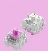 Two mechanical keyboard switches are shown against a light pink background. One switch is transparent with a pink stem, and the other is mostly white with gold pins. Produktbild-Vorschau