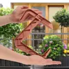 Hands holding a terracotta-colored balcony planter bracket with a blurred background of a balcony with plants and windows. Náhľad obrázku produktu