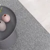 A close-up view of a gray rug next to a light wood floor, with a gray round table holding two small vases. Produktbild-Vorschau