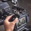 A hand is holding a small black wireless microphone transmitter in front of a camera's monitor, which displays two people talking. Náhľad obrázku produktu