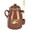 A brown ceramic jar with a lid and a wooden utensil holder attached, containing wooden tongs. The jar has an oval emblem with vegetables and text. Náhled obrázku produktu