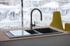 A modern kitchen sink with a black faucet and a glass cutting board, set against a window with a snowy cityscape view and a yellow chair in the background. Podgląd zdjęcia produktu