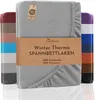 A folded gray fleece fitted sheet is displayed prominently, with other colored sheets stacked behind it. A brown paper band with product information is wrapped around the main sheet. Produktbild-Vorschau