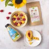 A flat lay of a breakfast bowl with fruit and nut butter, a bottle of nut butter, and a bag of cereal, with a sunflower in the background. Podgląd zdjęcia produktu