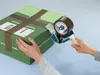 Hands are shown taping a green box shut with a tape dispenser. Produktbild-Vorschau