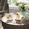 Two clear glass picture frames with abstract art are displayed on a wooden table next to a vase of white flowers and a mug. Podgląd zdjęcia produktu