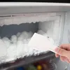 A hand is using a white plastic scraper to remove ice from inside a freezer. Produktbild-Vorschau