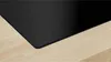A close-up view of the corner of a black induction hob resting on a light wood surface. Voir les images du produit