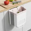 A white and gray foldable trash can with a pink liner is hanging from a kitchen cabinet. A cutting board with a tomato, peppers, and broccoli is on the counter above. Produktbild-Vorschau