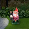 A garden gnome holding a lantern stands on a mushroom base in a garden setting. Produktbild-Vorschau