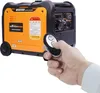 A hand is holding a remote control and pointing it towards an orange and black inverter generator. Produktbild-Vorschau