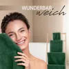 A woman is holding a dark green towel to her face in a bathroom setting, with a stack of dark green towels on a wooden rack in the background. Produktbild-Vorschau