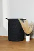 A dark gray woven laundry basket with brown handles stands on a wooden floor next to a white vase with dried pampas grass. Produktbild-Vorschau