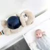 A plush otter toy with a blue sphere rests on a white surface next to a baby in a sleep sack. Produktbild-Vorschau