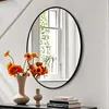 A mirror with a black frame is hung on a white wall, reflecting a window with plants. In front of the mirror, on a dark surface, are two vases with orange flowers and a stack of books. Podgląd zdjęcia produktu