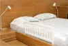 A white bed rail is placed on a bed with wooden headboard and nightstand. Voir les images du produit
