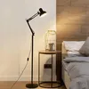 A black floor lamp is positioned next to a bedside table with a decorative cage on it, in a bedroom setting. Náhľad obrázku produktu