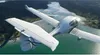 A white twin-engine airplane flying over a body of water and a forested coastline. Náhľad obrázku produktu