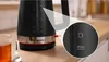 Close-up of a black electric kettle with a silver and red base, placed on a wooden countertop. A glass mug of tea is visible to the left. Anteprima dell’immagine prodotto