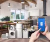 A collection of kitchen appliances arranged on a wooden surface in front of a blurred kitchen background, with a hand holding a smartphone displaying a control interface. Produktbild-Vorschau