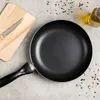 A frying pan, cutting board with a knife, and a bottle of oil are arranged on a textured gray surface, with scattered peppercorns. Produktbild-Vorschau