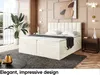 A bedroom scene featuring a cream-colored boxspring bed with a mattress and topper, set against a beige paneled wall. A large potted plant is visible to the left, and a dark wooden nightstand with decorative items is to the right. The overall ambiance is e Produktbild-Vorschau