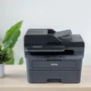 A Brother printer is placed on a wooden desk with a plant in the background. Produktbild-Vorschau