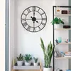 A large metal wall clock with Roman numerals is mounted on a white wall next to a shelving unit filled with plants and decorative items. Produktbild-Vorschau