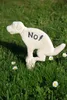 A white dog statue with 'No!' written on its side, placed in a grassy area with daisies. Produktbild-Vorschau