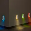 Four wall-mounted LED lights with different colored glows (blue, yellow, green, red) illuminating a dark hallway or room. Náhled obrázku produktu