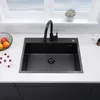 A black kitchen sink with a black faucet is shown on a white countertop, with a window and a plant in the background. Náhled obrázku produktu