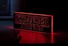 A Stranger Things logo light is displayed on a dark surface, with a hint of a computer tower in the background. Produktbild-Vorschau