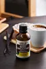 A bottle of vanilla extract is placed on a wooden surface next to a cup of coffee and vanilla beans. Náhľad obrázku produktu