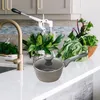 A granite saucepan with a glass lid sits on a white countertop in a kitchen setting, with fresh vegetables and a faucet in the background. Produktbild-Vorschau