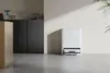 A white robot vacuum cleaner with its charging dock is placed on a gray floor in front of a light-colored wall. To the left, a dark cabinet with some decorative items is visible. Náhled obrázku produktu