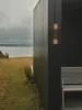 A dark-colored building with a wooden wall light fixture and a wooden bench on the side, with a body of water and a cloudy sky in the background. Produktbild-Vorschau
