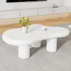 A white, irregularly shaped coffee table with two cylindrical legs sits on a gray rug in a living room setting. A TV stand is visible in the background. Produktbild-Vorschau