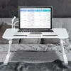A white laptop table with a laptop and a small lamp is set up on a bed with gray bedding. Produktbild-Vorschau