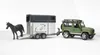 A toy horse trailer being towed by a toy green Land Rover Defender, with a toy black horse standing on the trailer ramp. Produktbild-Vorschau