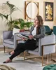 A woman is sitting in a gray velvet armchair with a gold metal frame, reading a magazine. The room is decorated with plants and artwork. Náhled obrázku produktu