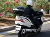 A person riding a white scooter with a black top case on a road with greenery and flowers. Anteprima dell’immagine prodotto
