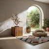 A rustic wooden column with a decorative branch sculpture sits next to an arched doorway opening to a garden. A patterned rug and floor cushions are in the foreground. Náhled obrázku produktu